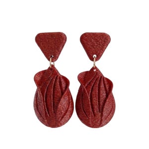 Peut inclure: Une paire de boucles d'oreilles pendantes rouge foncé composées d'un clou triangulaire relié à un pendentif plus grand en forme de pétale. La surface présente une texture brodée fine et détaillée avec des stries verticales sur l'ensemble.