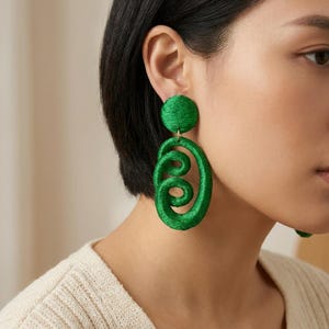 Può includere: Un paio di orecchini verde brillante con un perno circolare collegato a un componente ovale pendente più grande. La superficie degli orecchini è rivestita da un materiale testurizzato simile a un filo che forma un motivo a spirale continuo.