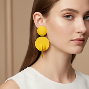 May include: A bright yellow statement earring featuring a small textured circular stud connected to a larger flat circular disc. A vertical semi-circular piece is attached to the centre of the bottom disc, creating a three-dimensional geometric shape.