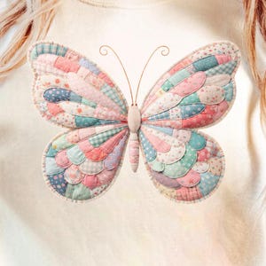 May include: Cream-colored t-shirt featuring a large, colorful butterfly design. The butterfly is made of patchwork fabric in pastel shades of pink, blue, and green. The butterfly has delicate antennae and a textured appearance.