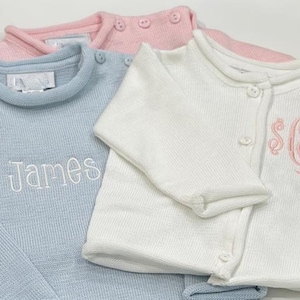 May include: Close-up of three baby sweaters in pastel colors. A light blue sweater with the name "James" embroidered in white. A white cardigan with pink monogram. A pink sweater is visible in the background.