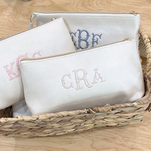 May include: Three off-white zippered pouches with gold-tone zippers and embroidered initials in pink, blue, and beige. The pouches are arranged in a woven basket, suggesting a gift set or storage solution.