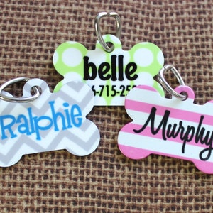 May include: Three dog bone-shaped ID tags with different patterns and names. The first tag is green with white polka dots and the name "belle" in black. The second tag is gray with a chevron pattern and the name "Ralphie" in blue. The third tag is pink with white stripes and the name "Murphy" in black.