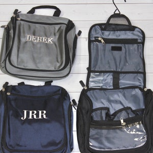 May include: A collection of travel toiletry bags in various colors. One gray bag has "DEREK" embroidered on it. A navy blue bag displays "JRR". A black bag is open, revealing multiple compartments and a hanging hook. All bags have zippers.