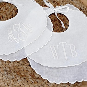 May include: Two white baby bibs with scalloped edges and embroidered initials. One bib features the initials "LSM", and the other has "WTB". Each bib has a white ribbon tie at the neck, perfect for a baby shower gift.