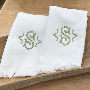 May include: Two white rectangular hand towels featuring a light green embroidered monogram of the letter S in a decorative script font. Each towel has a fringed bottom edge and a smooth fabric texture, presented as a matching pair of linens.