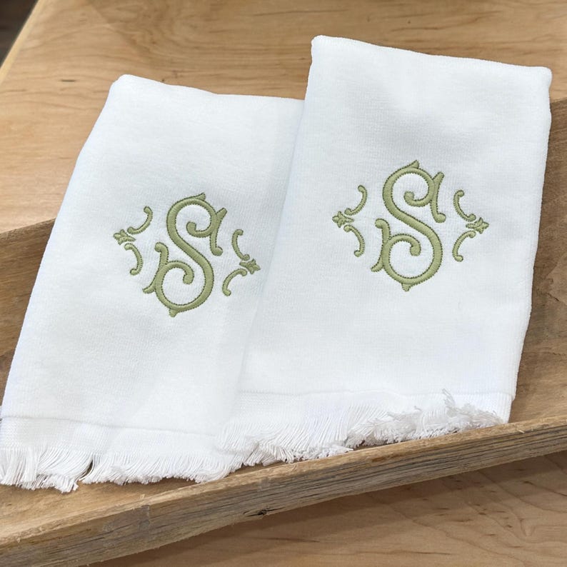 May include: Two white rectangular hand towels featuring a light green embroidered monogram of the letter S in a decorative script font. Each towel has a fringed bottom edge and a smooth fabric texture, presented as a matching pair of linens.