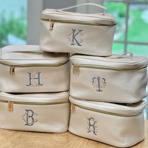 May include: Cream-colored cosmetic bags with gold zippers and handles. Each bag features a different embroidered initial in a blue-gray color. The bags are rectangular with a structured design, suitable for travel or storage.