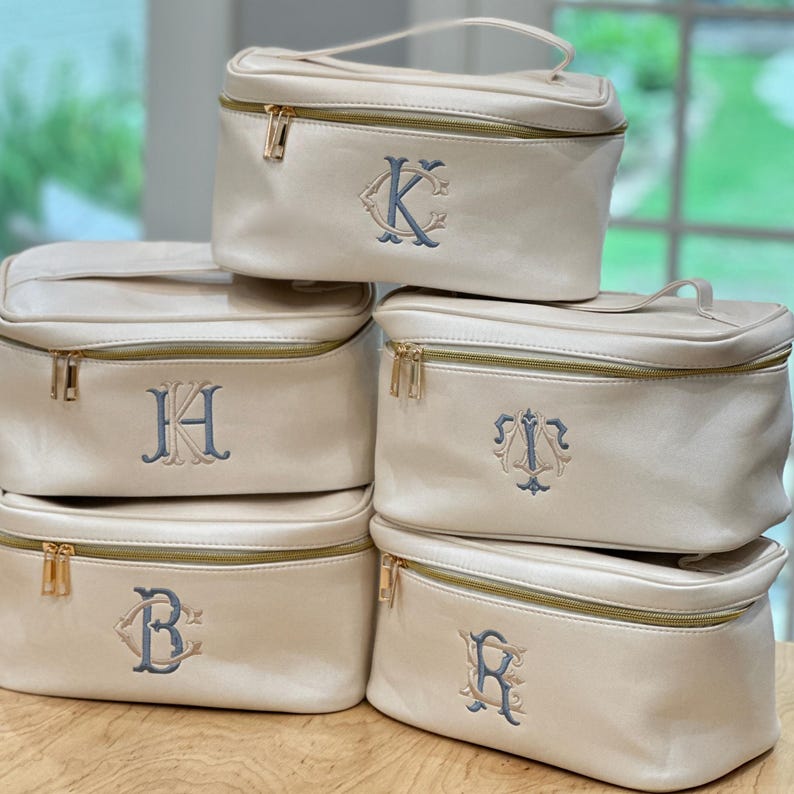 May include: Cream-colored cosmetic bags with gold zippers and handles. Each bag features a different embroidered initial in a blue-gray color. The bags are rectangular with a structured design, suitable for travel or storage.