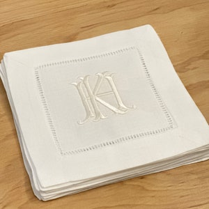 May include: A stack of white linen napkins with a white embroidered monogram "JK" in a script font.