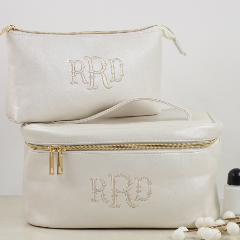 May include: Two ivory-colored cosmetic bags, one rectangular and one smaller, are stacked. Both bags have gold zippers and embroidered initials. The larger bag has a handle. The bags are on a white surface.