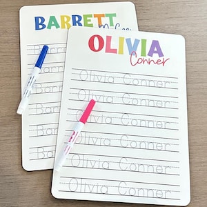 May include: Two white dry-erase boards with colorful names at the top. The top board says "BARRETT" in rainbow letters. The bottom board says "OLIVIA Conner" in rainbow letters. Both boards have lined spaces for writing practice.