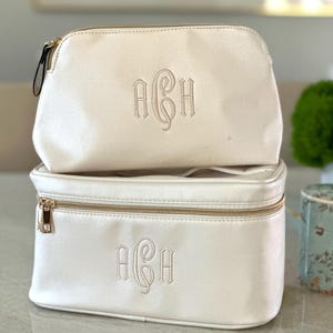May include: Two ivory-colored cosmetic bags stacked on top of each other. Each bag has a gold zipper and a monogram in the center. The top bag is smaller and rounded, while the bottom bag is rectangular.