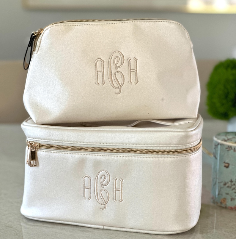 May include: Two ivory-colored cosmetic bags stacked on top of each other. Each bag has a gold zipper and a monogram in the center. The top bag is smaller and rounded, while the bottom bag is rectangular.