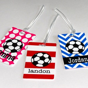 May include: Three personalized luggage tags with soccer ball designs. The tags are pink with white polka dots, red with white stripes, and blue with white chevron patterns. Each tag has a black soccer ball and a name: Emma, Landon, and Jordan.
