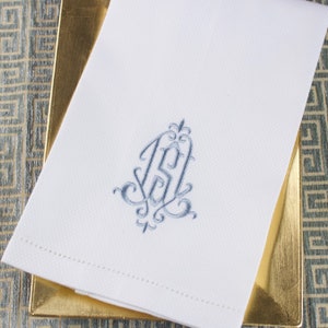 May include: White linen towel with a light blue embroidered monogram. The monogram features ornate script initials and decorative flourishes. The towel is folded and placed on a gold-colored tray, with a patterned background.
