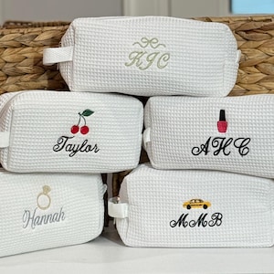 May include: Five white waffle-textured cosmetic bags with embroidered designs. One bag has the initials "KJC", another has cherries and the name "Taylor", a third has a nail polish bottle and the initials "AHC", a fourth has a ring and the name "Hannah", and the last has a yellow car and the initials "MmB".