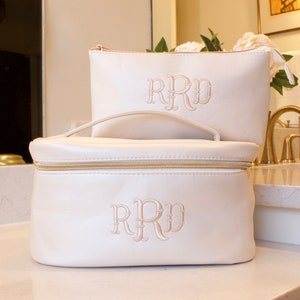 May include: Two white toiletry bags with gold zippers and a monogram of "RRD" in a script font. The larger bag has a handle.