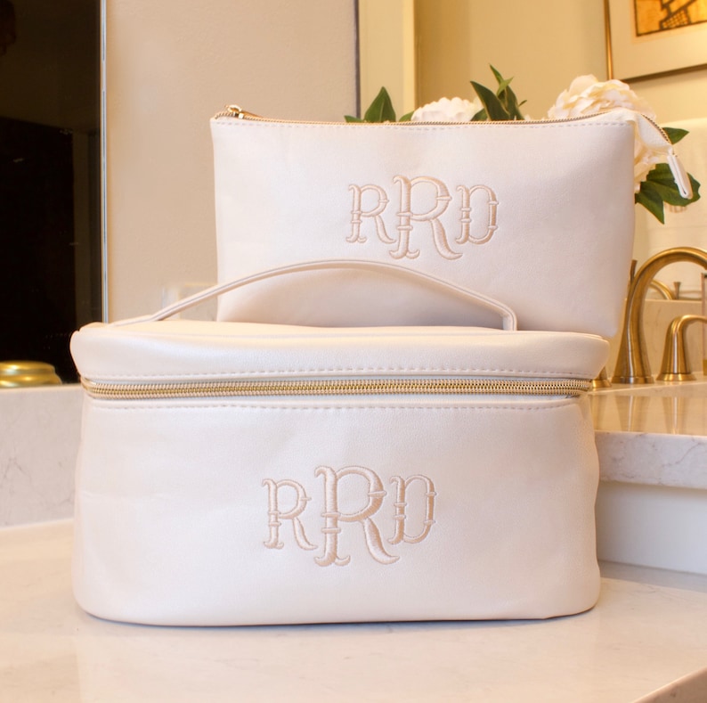 May include: Two ivory-colored cosmetic bags with gold zippers and embroidered initials. The larger bag has a handle and a rectangular shape, while the smaller bag is rectangular. The initials "RRD" are embroidered on both bags.