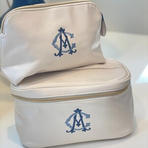 May include: Two ivory-colored cosmetic bags, one rectangular and one rounded, stacked on top of each other. Each bag features a blue embroidered monogram and a gold zipper. The bags are made of a smooth, leather-like material.