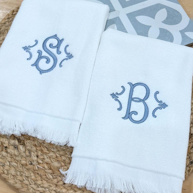 May include: Two white rectangular towels featuring fringed edges at the bottom. Each towel is embroidered with a single large letter in a dusty blue thread, one displaying the letter S and the other displaying the letter B, both with scrollwork.