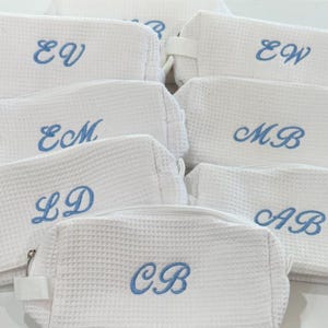 May include: A set of white waffle-textured zippered pouches with blue embroidered initials. The initials include LD, CB, AB, MB, ECM, EW, and EV.