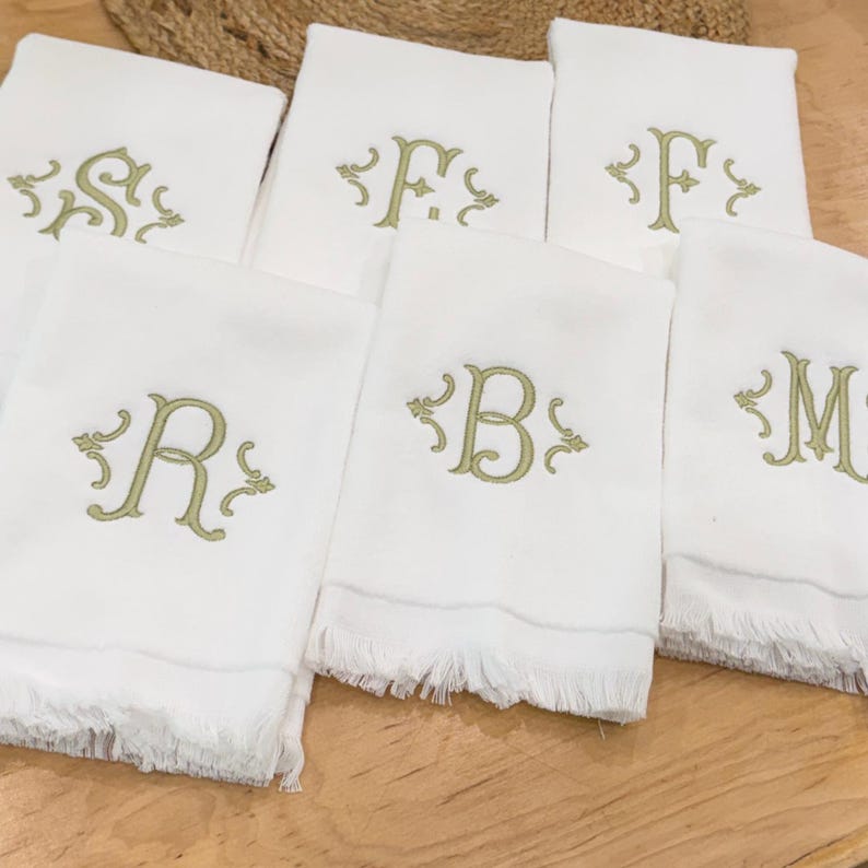 May include: Six white rectangular hand towels with fringed edges, each featuring a centered embroidered letter in a light olive green thread. The letters S, F, F, R, B, and M are surrounded by decorative scrollwork accents in the same green color.