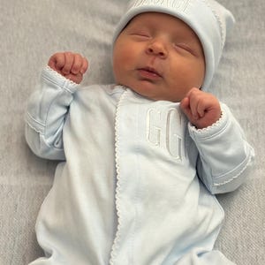 May include: A newborn baby wearing a light blue onesie and matching cap. The cap is embroidered with the name "GEORGE" and the onesie has the initials "GG". The outfit has white scalloped trim.