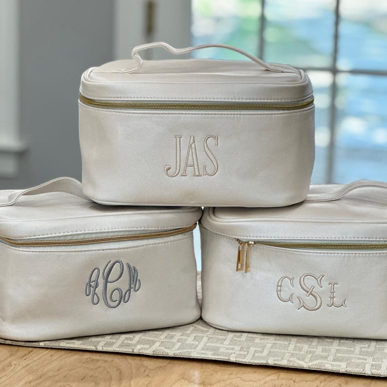 May include: Three ivory-colored cosmetic bags stacked on top of each other. Each bag has a gold zipper and a handle. The top bag has the embroidered letters "JAS", the bottom left bag has "ACD", and the bottom right bag has "CSL".