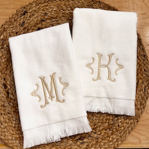 May include: Two white hand towels with fringed edges. The towels are embroidered with the letters "M" and "K" in a gold script font.