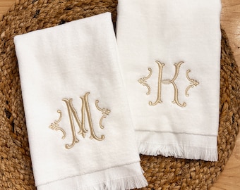 May include: Two white hand towels with fringed edges. The towels are embroidered with the letters "M" and "K" in a gold script font.