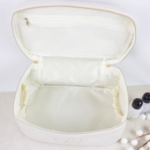 May include: Open white cosmetic bag with a zippered top and interior mesh pocket. The bag is made of a smooth, light-colored material and has a zippered closure. The interior is lined with a light-colored fabric. The bag has the initials "RPR" embroidered on the front.
