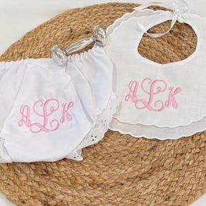 May include: A white fabric diaper cover and a matching white bib, both featuring pink embroidered monogram letters ALK. The bib has a scalloped edge and a white ribbon tie. A silver-toned metal rattle is positioned between the two items.