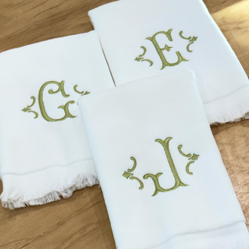 May include: Three white rectangular towels featuring decorative fringe on the bottom edges. Each towel is embroidered with a single large letter in a light olive green thread, specifically the letters G, E, and J, surrounded by ornate scrollwork.