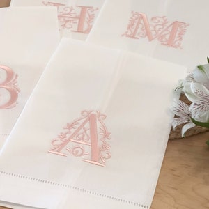 May include: White linen towels with embroidered pink floral initial letters. The letter "A" is prominently displayed on the front towel. Other towels with the letters "B", "H", and "M" are also visible. The towels are neatly arranged on a wooden surface.