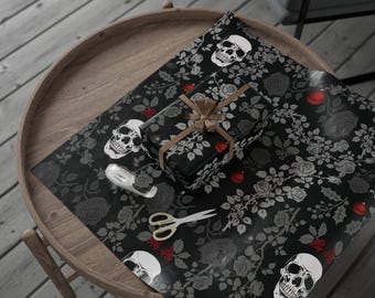 May include: A wrapped gift with a brown ribbon sits on black wrapping paper with a skull and rose pattern. A roll of tape and scissors are also on the paper, which is on a wooden table.