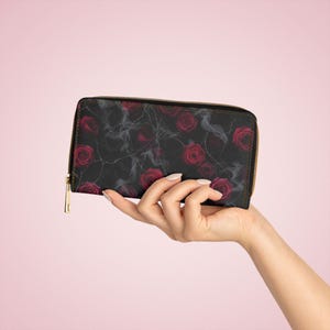 May include: Black wallet with a gold-coloured zip, patterned with red roses and wispy grey accents. The wallet is held in a hand against a pink background. The wallet is rectangular and has a zip-around closure.