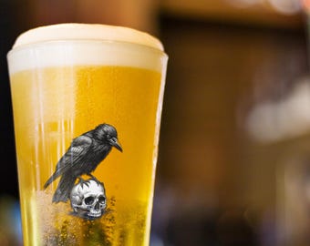 May include: A tall glass of beer with a thick, foamy head. A black raven is perched on a skull graphic on the glass. The beer is a golden color, and the background is blurred.