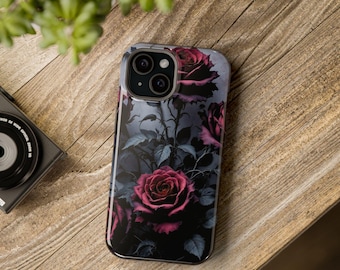 May include: A smartphone case featuring a dark floral design with deep red roses and gray leaves against a dark background. The case is on a wooden surface, with a camera and plant in the background.