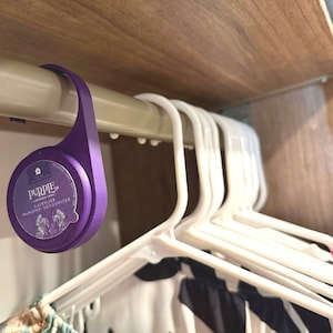 Könnte beinhalten: Ein lila Hängedeodorant mit dem Wort "PURPLE" auf der Vorderseite, das an einer Kleiderstange hängt. Das Deodorant ist rund mit einem gebogenen Haken. Weiße Kleiderbügel und Kleidung sind im Hintergrund zu sehen.