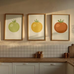 May include: Three framed tomato art prints in a kitchen setting. Each print features a different colored tomato: green, yellow, and orange. The frames are a light brown color, and the background is a neutral white.