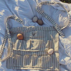 May include: A striped egg gathering apron with multiple pockets and a neck strap. The apron is made of a light-colored fabric with vertical stripes and a small patch with a chicken illustration. Three eggs are visible.