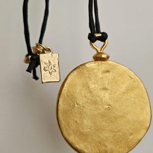 May include: Two gold-colored pendant necklaces. One features a rectangular charm with a floral design, the other a large, round pendant. Both are suspended from black cords. The pendants have a textured surface and are set against a neutral background.