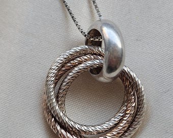 May include: A silver-toned necklace featuring a pendant composed of three interlocking circular rings. Two of the rings have a textured, diamond-cut surface, while the third, larger ring is smooth and polished, serving as the bail for the chain.