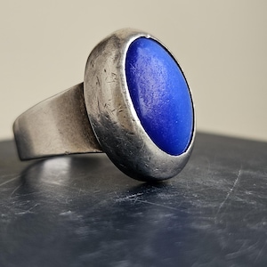 May include: A silver ring featuring a vibrant blue oval stone. The ring has a substantial band and a bezel setting. The ring is displayed against a dark surface.