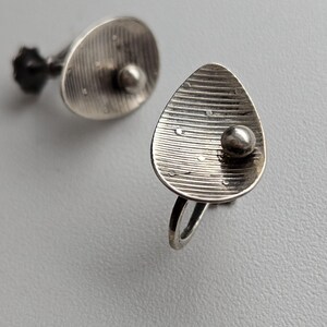 Peut inclure: Boucles d'oreilles argentées au design unique. Chaque boucle d'oreille présente un élément en forme de goutte d'eau avec des lignes horizontales et un petit accent rond. Les boucles d'oreilles ont une tige et une petite boule à l'arrière.