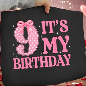 Peut inclure: Coussin noir avec le texte "IT'S MY BIRTHDAY" en rose. Le chiffre 9 est rose avec des pois blancs et un nœud rose. Le coussin est tenu par une personne.