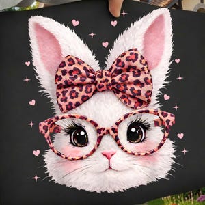 Peut inclure: Un carré noir avec un visage de lapin blanc mignon aux oreilles roses, portant un nœud et des lunettes à imprimé léopard. Le lapin a de longs cils et est entouré de cœurs et d'étoiles roses. L'image est tenue par une personne.
