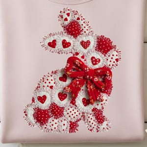 Peut inclure: Un sweat-shirt rose clair orné d'un motif de lapin en patchwork. Le lapin est fait de tissu rouge et blanc avec des motifs de cœurs et de pois, et un nœud floral rouge. Le sweat-shirt est plié sur une surface en bois blanc.