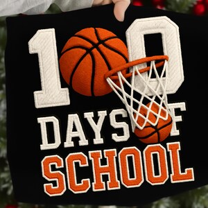 Puede incluir: Cuadrado negro con el texto "100 DÍAS DE ESCUELA" en blanco y naranja. El diseño incluye una pelota de baloncesto naranja y una red de baloncesto. La imagen se sostiene frente a un árbol de Navidad.